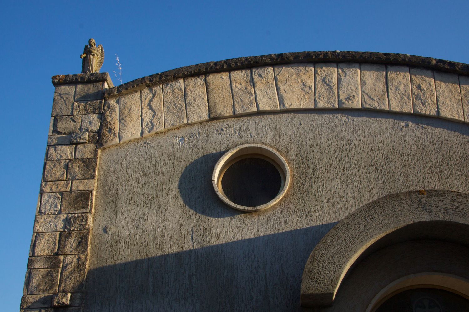 Stone church rooftop with circular oculus window and curved arch against clear blue sky.