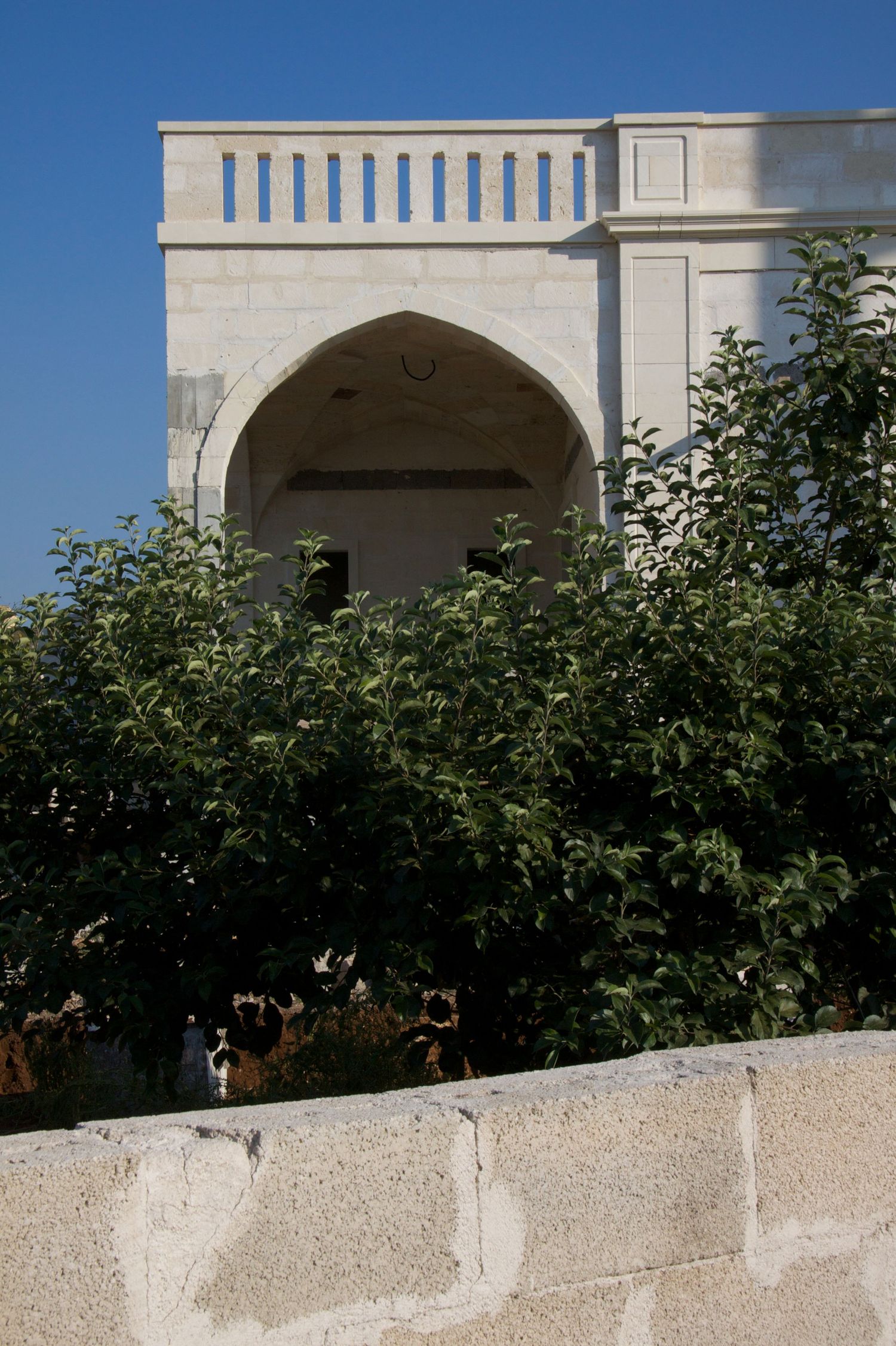 White stone building with a pointed arch entrance surrounded by lush green trees under a blue sky.