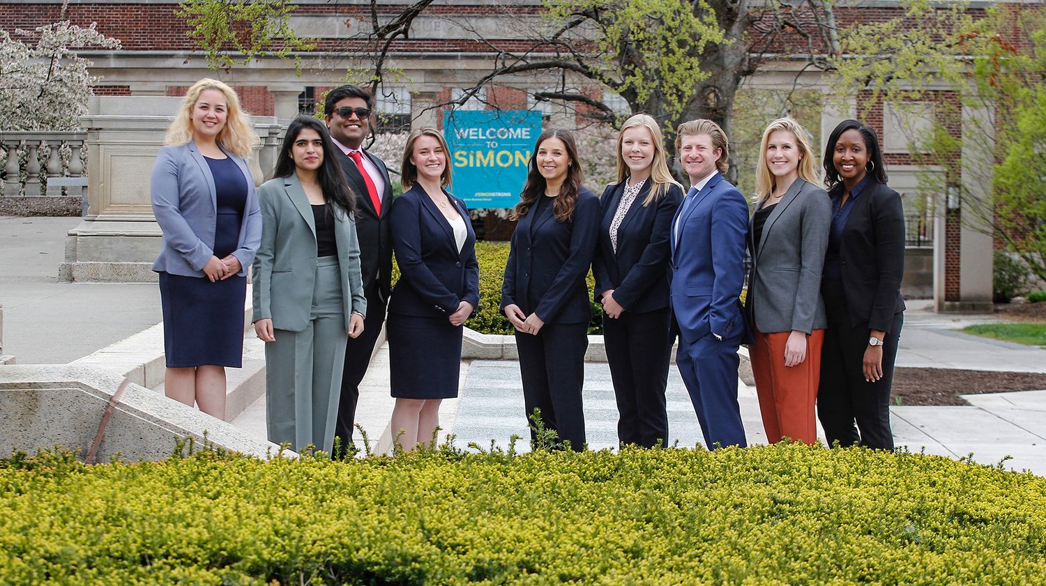 Group of nine professionally dressed women posing outdoors near a Simon welcome sign.