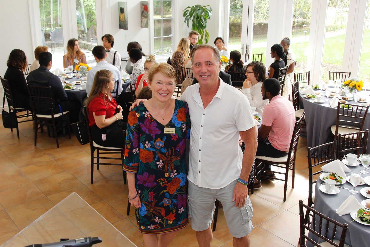 Two smiling guests posing together at an indoor university dining event with guests seated behind.