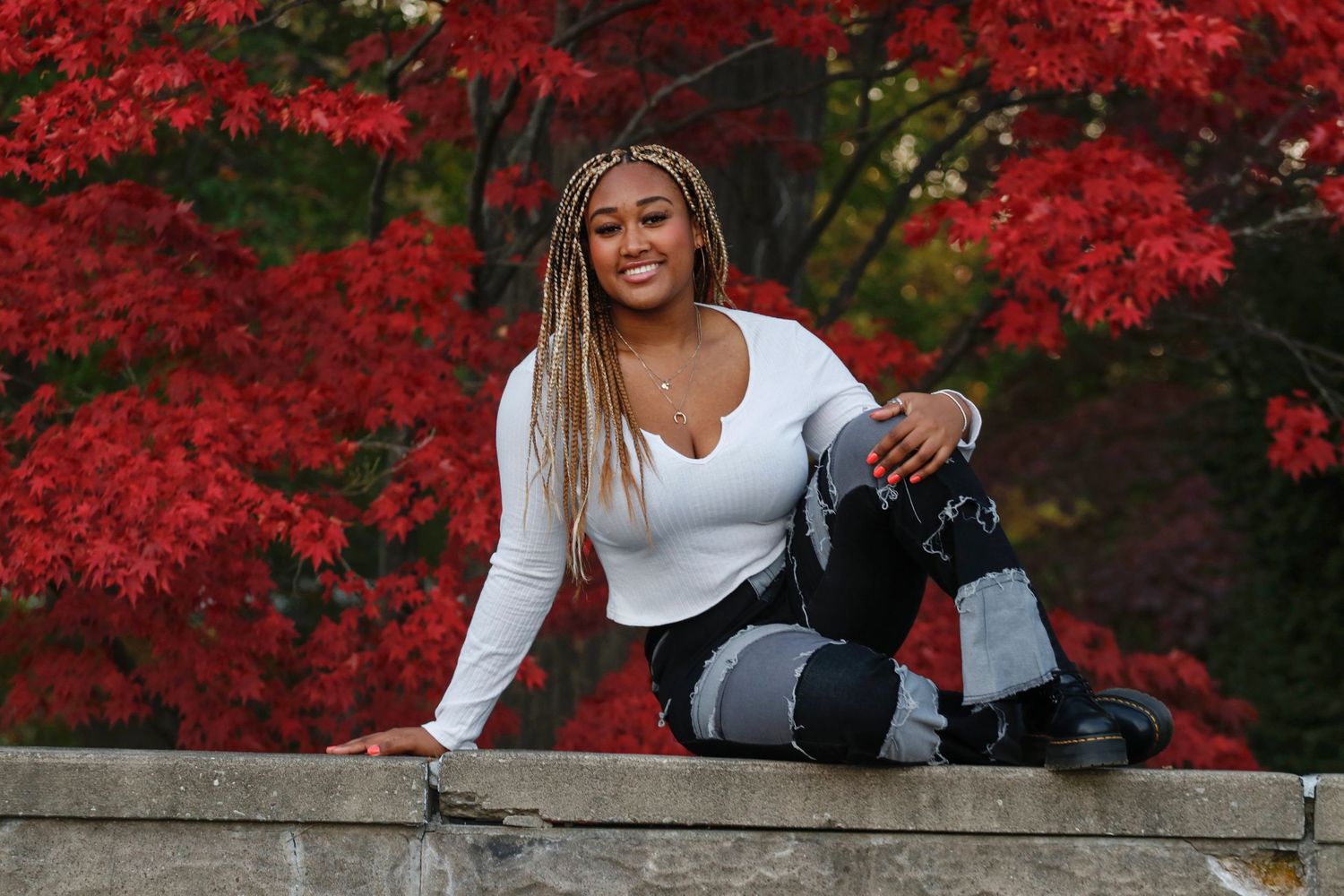 Young woman with braids posing confidently against vibrant red autumn foliage outdoors.