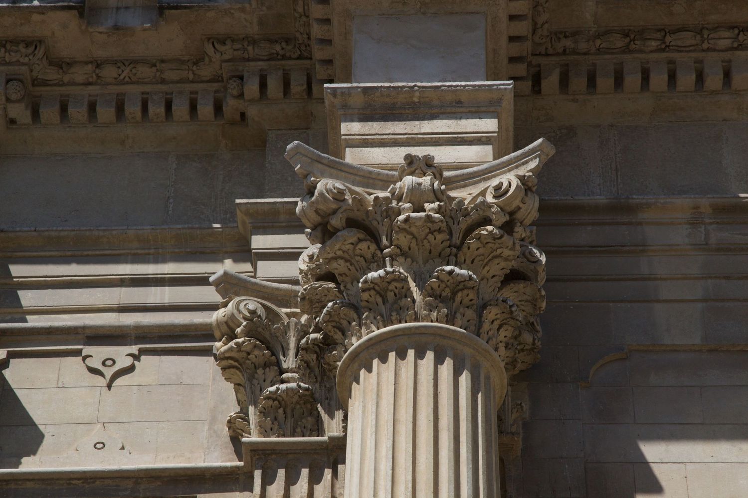Ornate Corinthian column capital with acanthus leaf carvings on a classical stone building facade in bright sunlight.