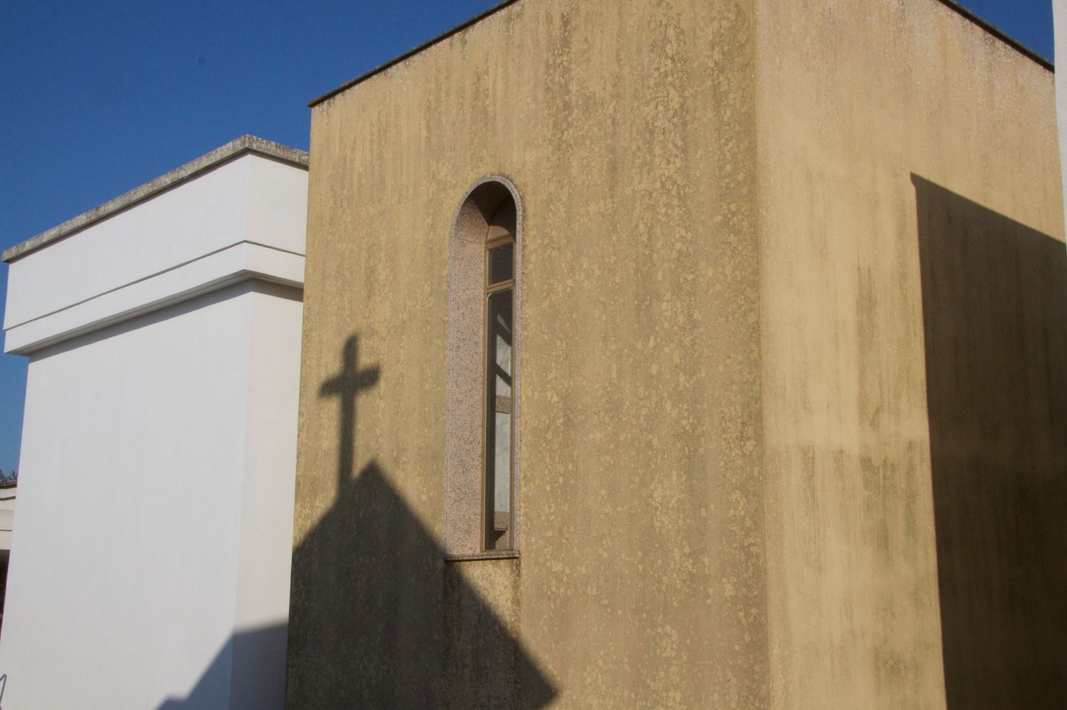 Shadow of a cross cast on a textured yellow stone church exterior wall under bright blue sky.