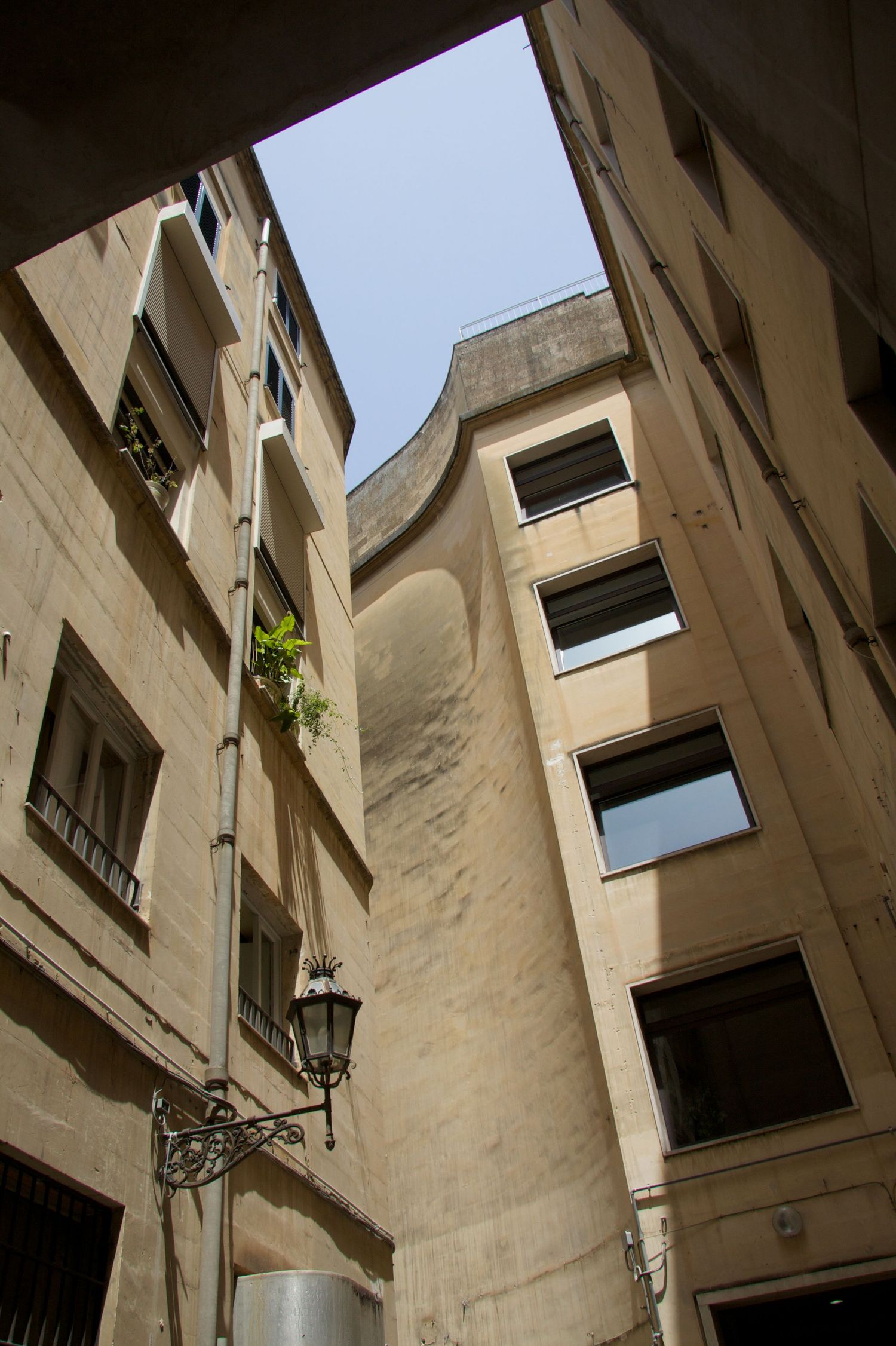 Narrow urban courtyard between tall beige stone buildings with a vintage street lamp and blue sky above.
