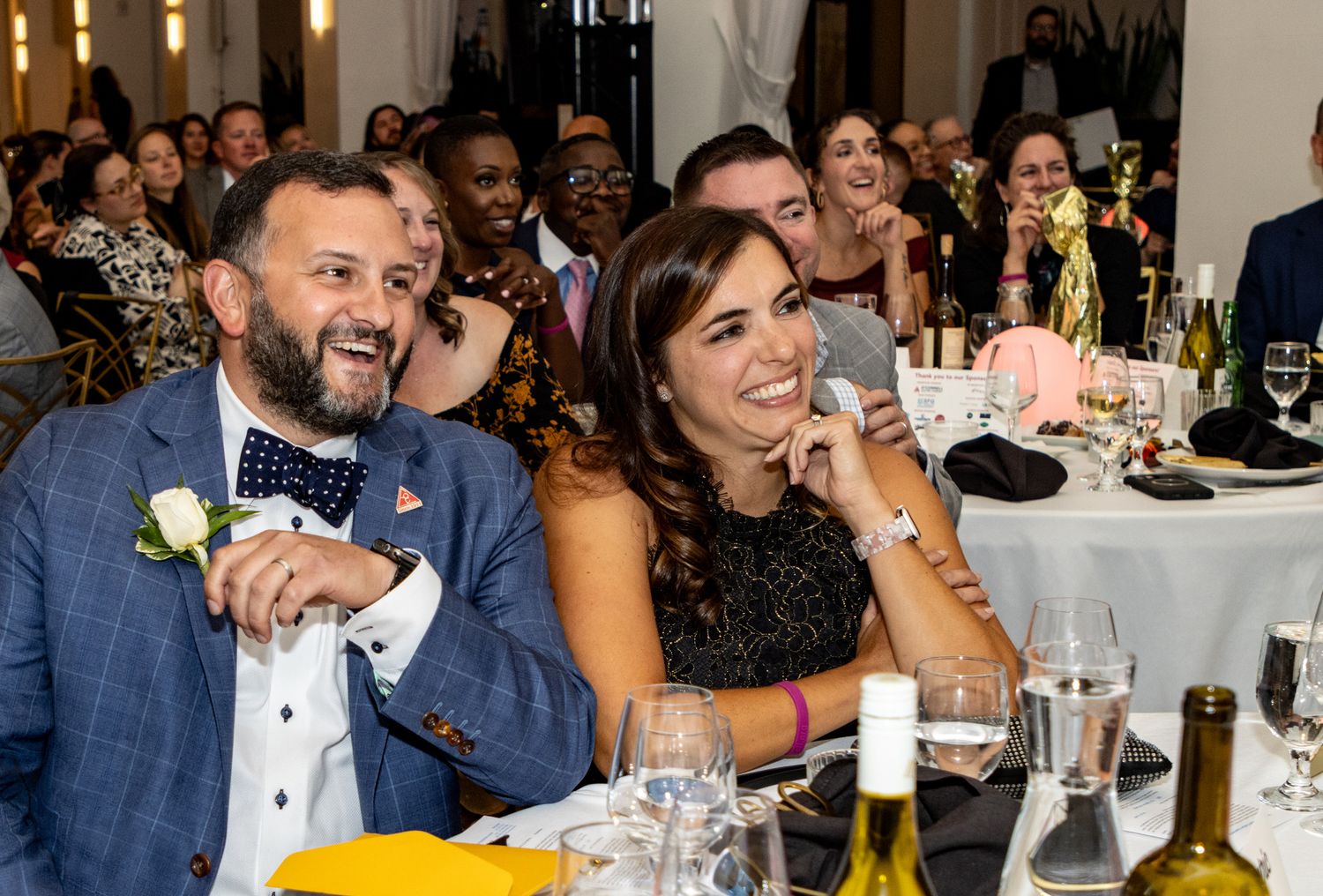 Two guests laugh and smile at a formal wedding reception dinner, seated at an elegantly set table with wine bottles.