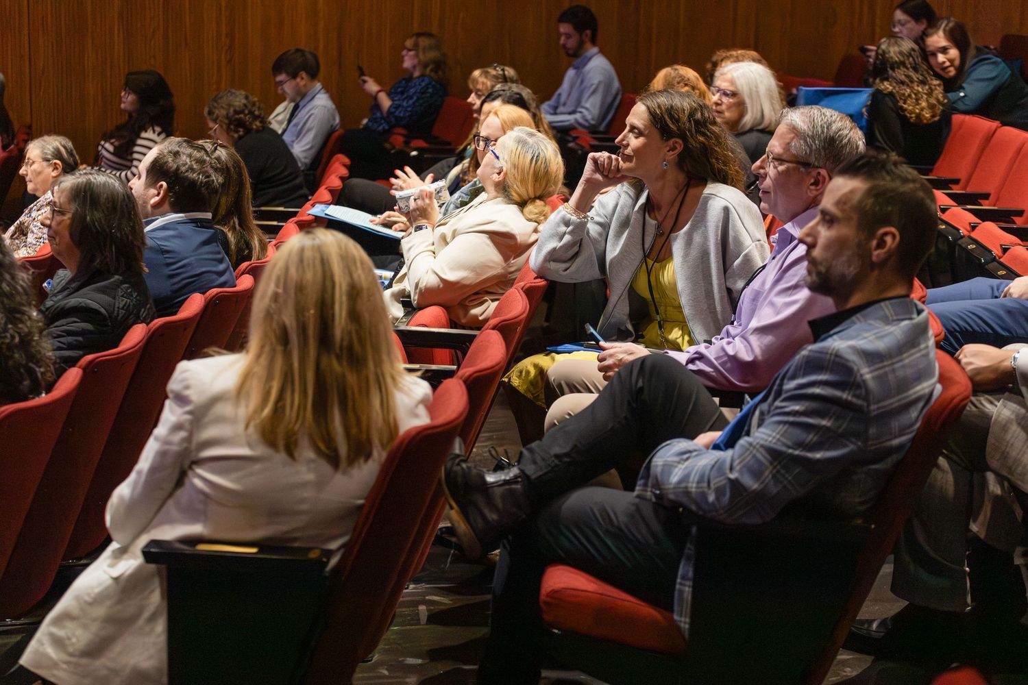 Audience members seated in red chairs at an indoor conference or theater presentation.