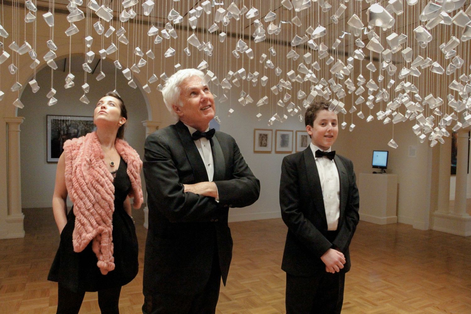 Three elegantly dressed guests admire hanging glass installations at an upscale art gallery event.