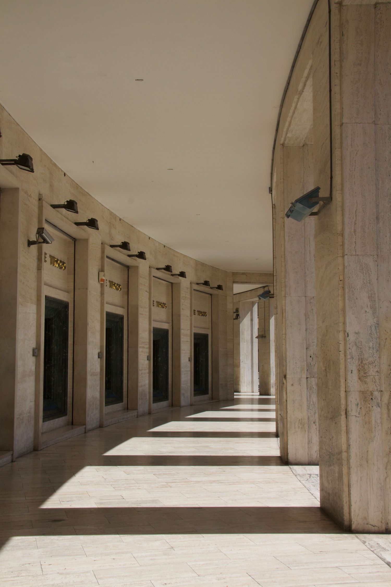 Curved modernist colonnade corridor with marble floors, wall-mounted lamps, and repeating doorways casting dramatic shadows.