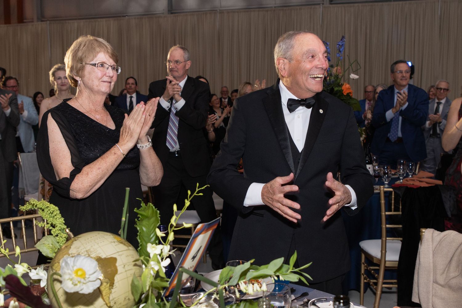 Elegantly dressed couple stands and smiles as guests applaud at a formal gala dinner event.