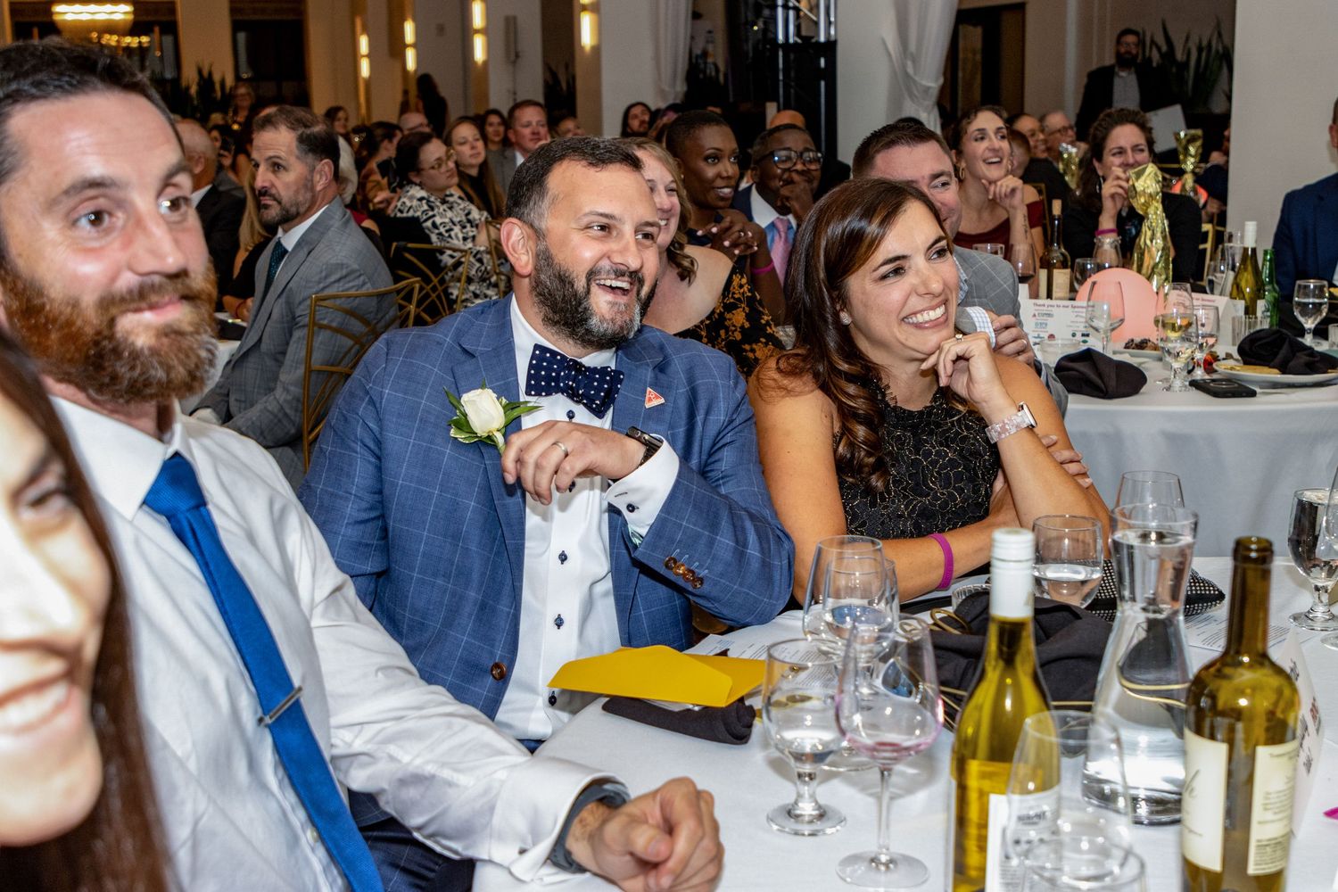 Guests laugh and celebrate together at a formal wedding reception dinner with wine and floral decor.