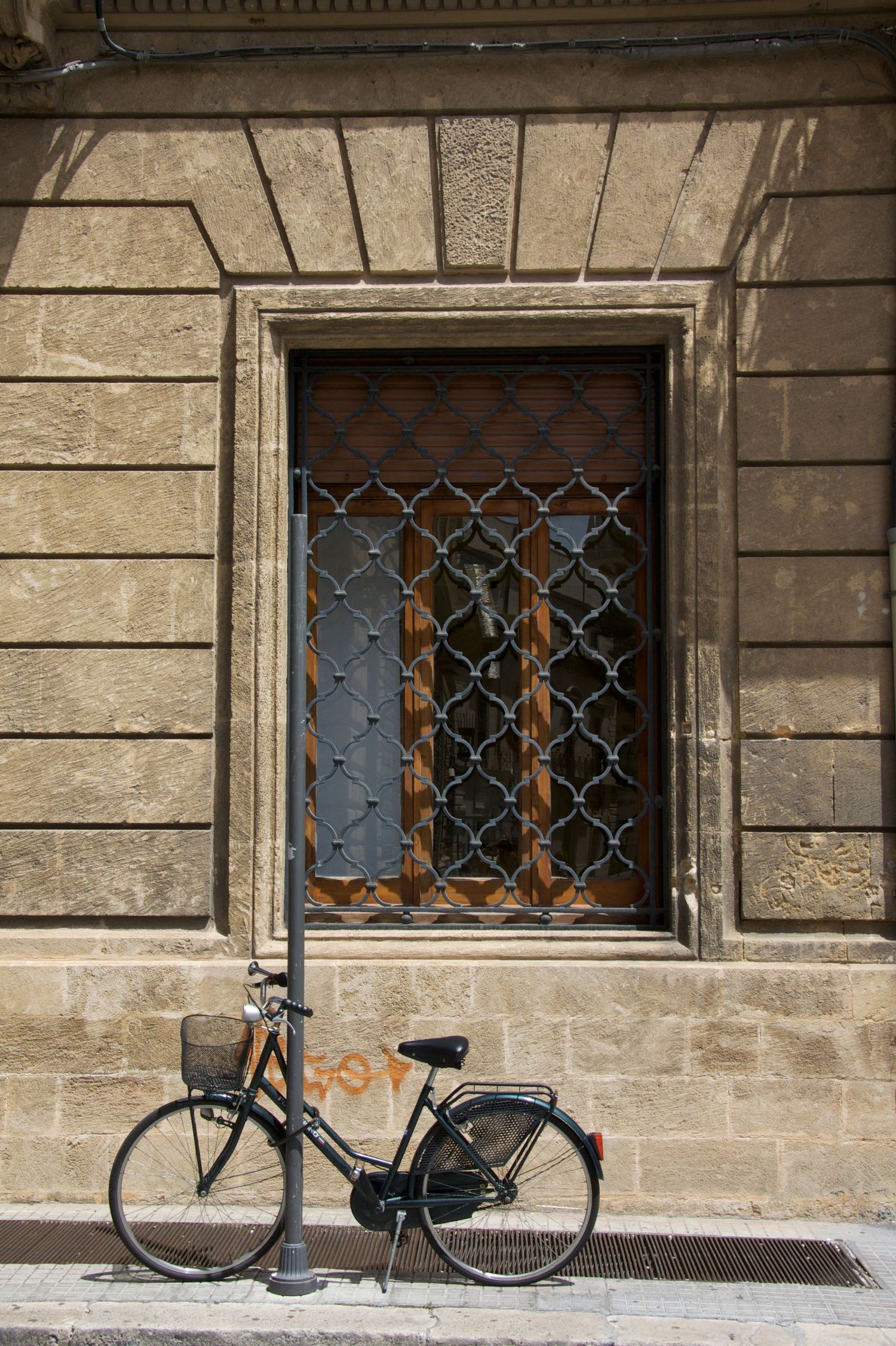 Black bicycle parked against historic stone building wall beside ornate iron-grilled window.