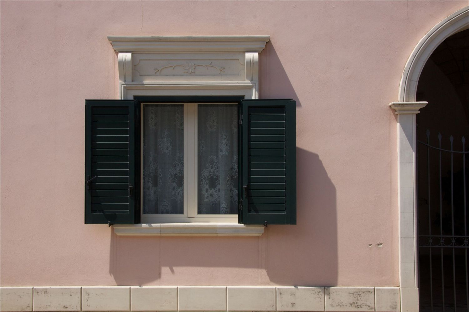 Pink stucco building exterior featuring a white-framed window with open dark green wooden shutters.