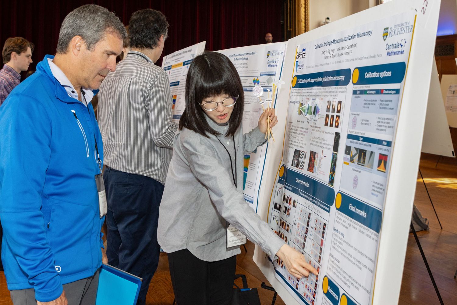 Woman presents research poster to male colleague at academic conference in a large auditorium.
