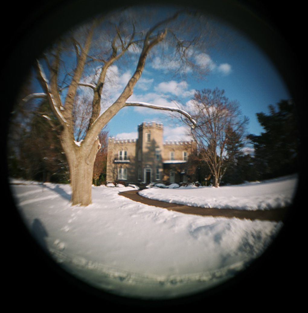 Fisheye lens photo of a snow-covered castle-like stone mansion with bare trees under a blue sky in winter.