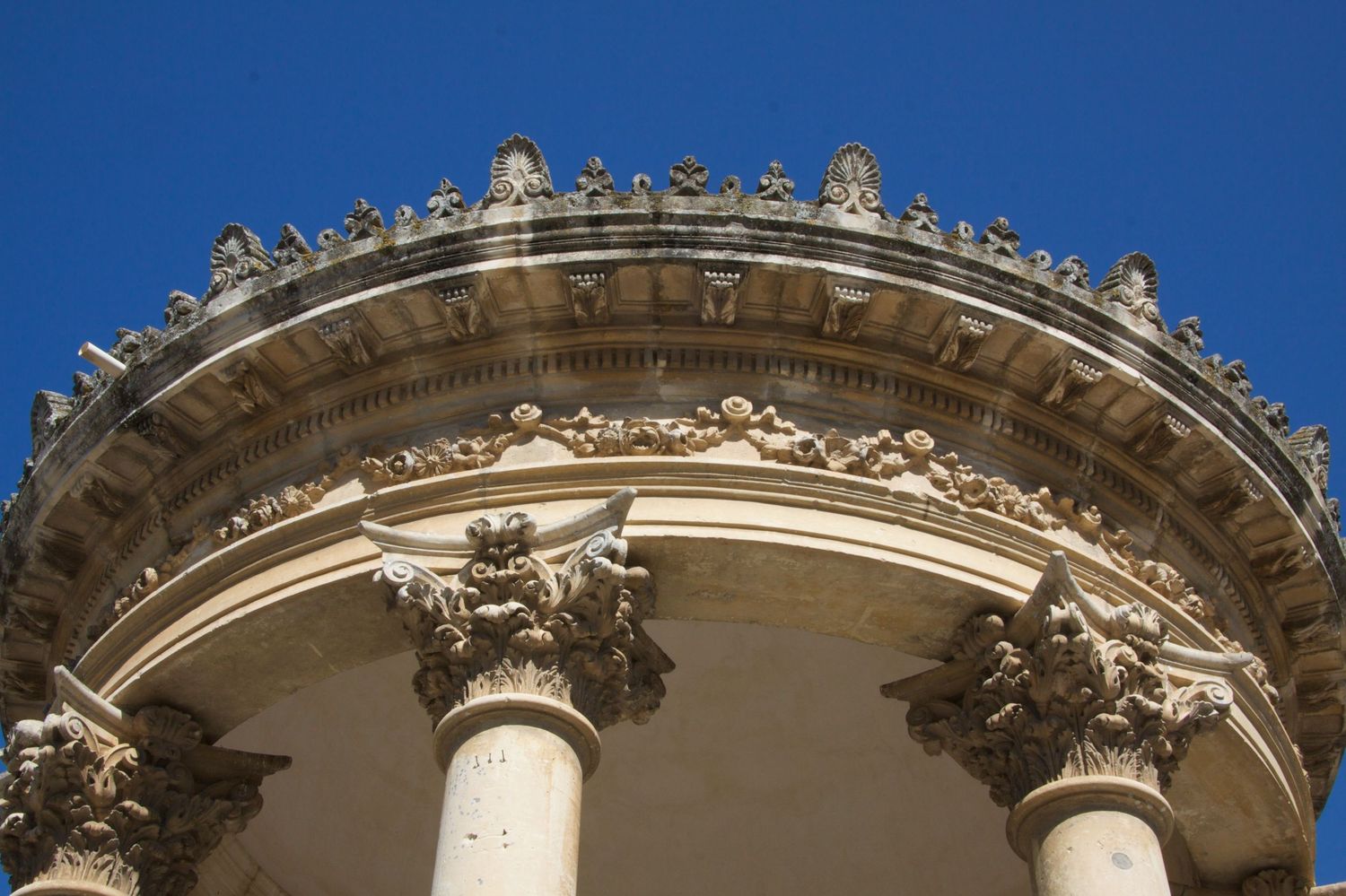 Ornate Baroque rotunda roofline with Corinthian columns, decorative friezes, and statues against a vivid blue sky.