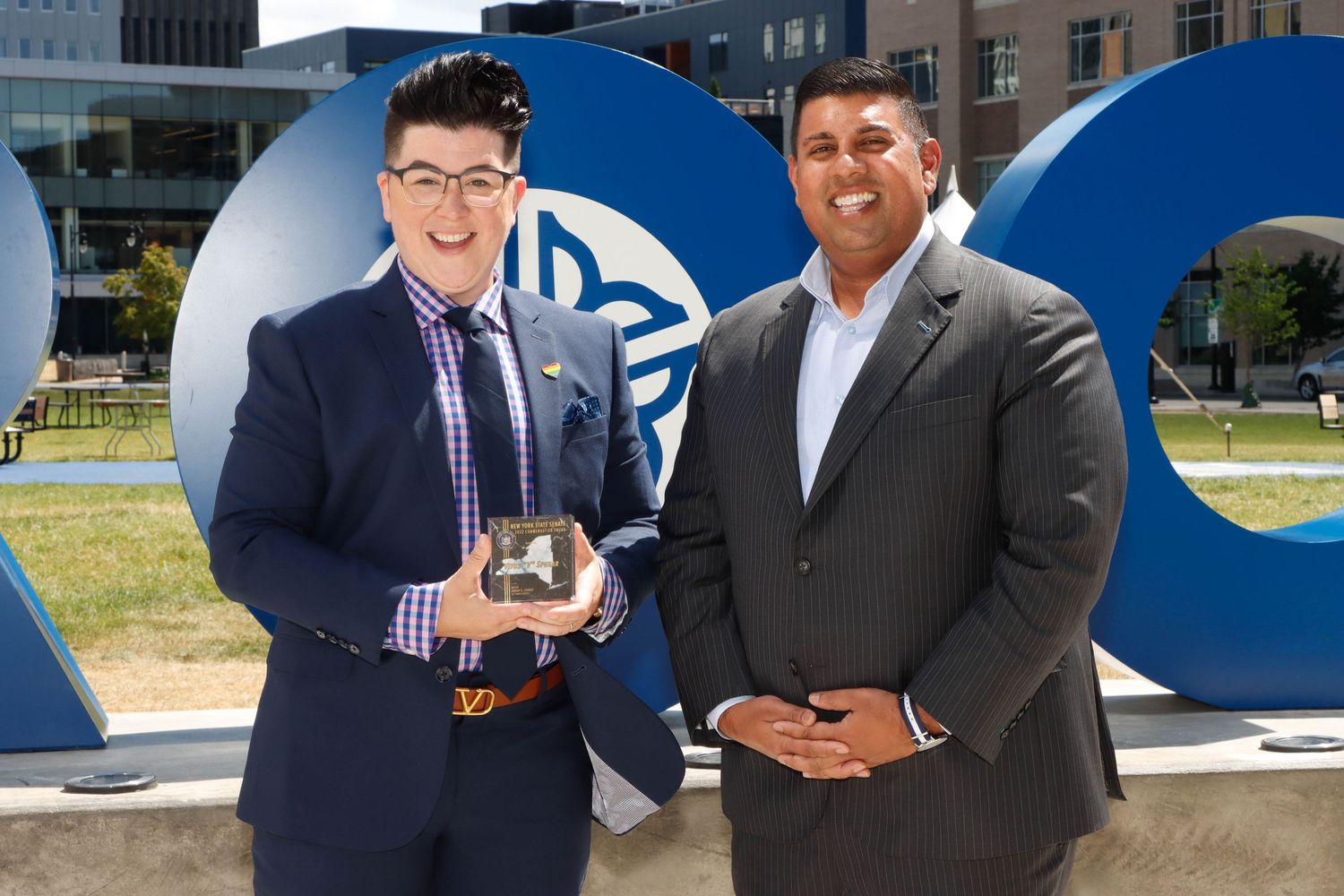 Two people posing with award in front of large blue circular sculpture outdoors.