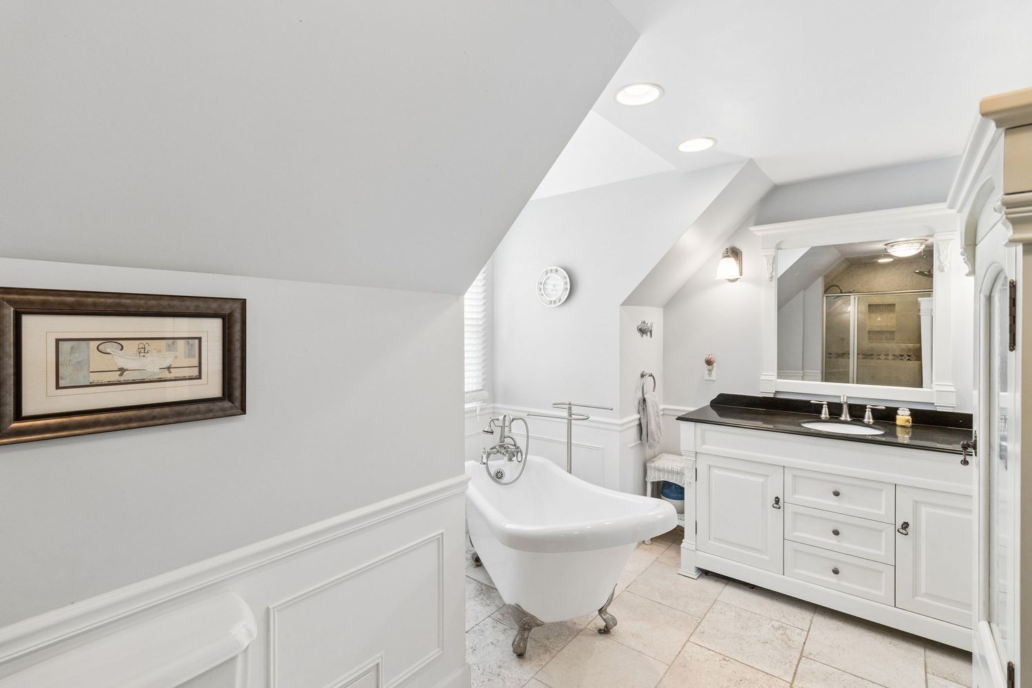 Elegant bathroom with clawfoot soaking tub, white wainscoting, vanity with dark countertop, and skylight.