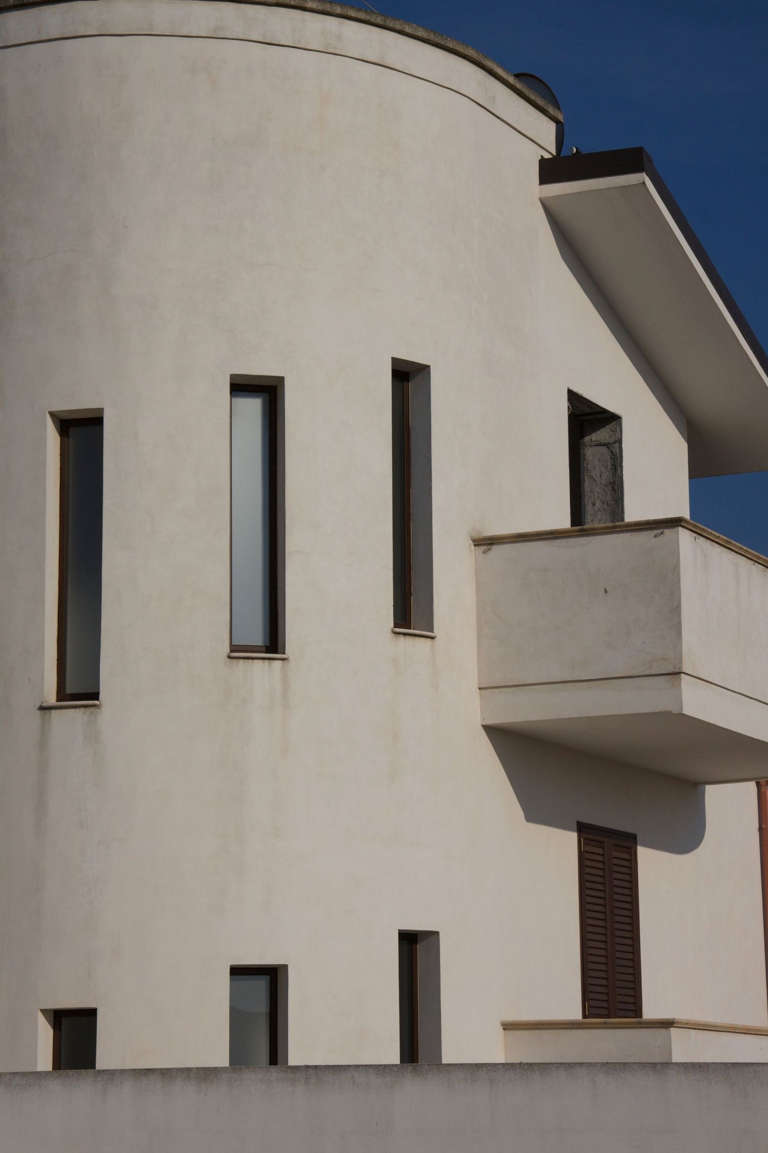Modern white stucco building with curved cylindrical tower, narrow vertical windows and balcony against clear blue sky.