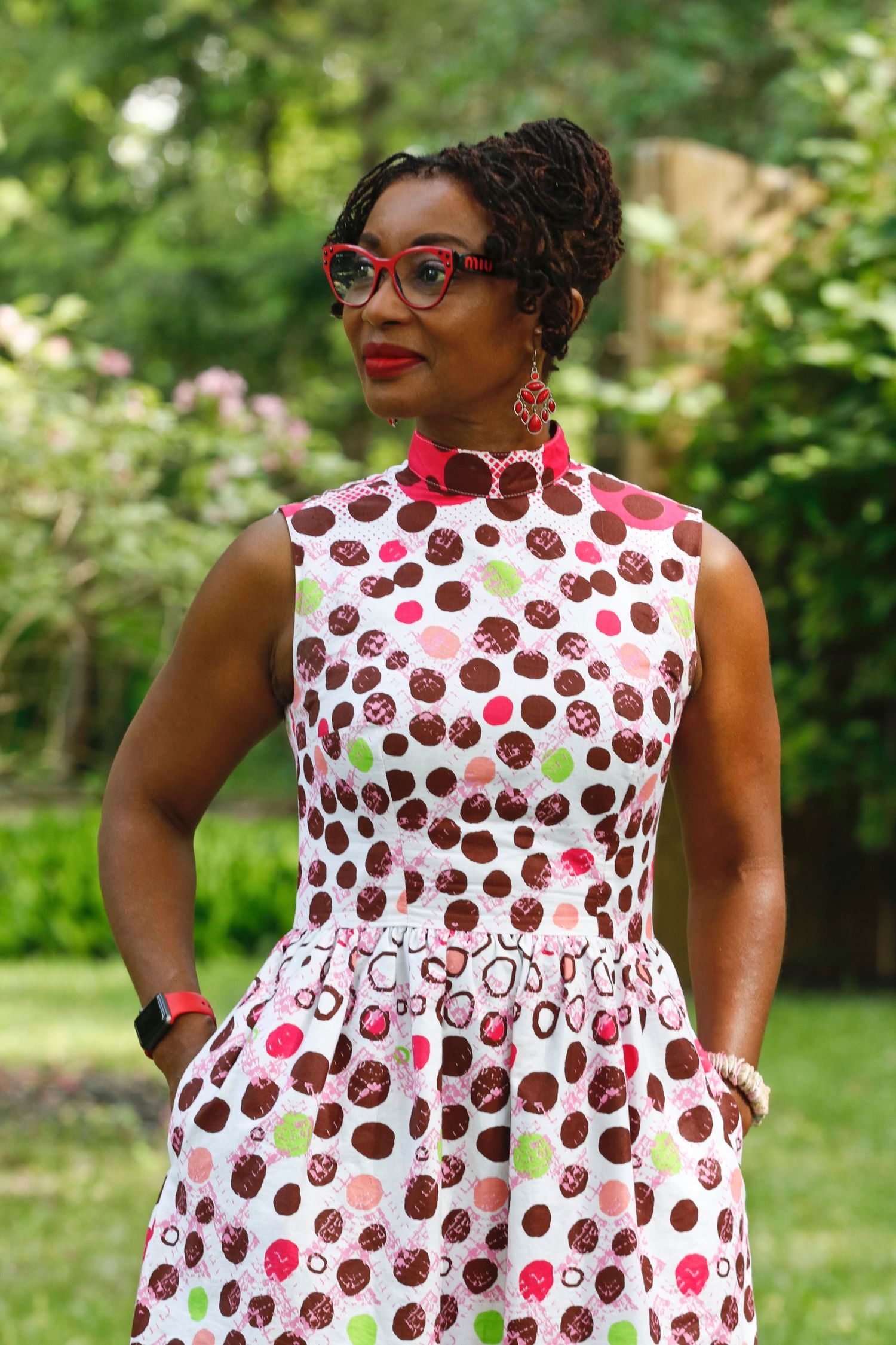 Woman in polka dot dress and red glasses posing confidently in a garden setting.
