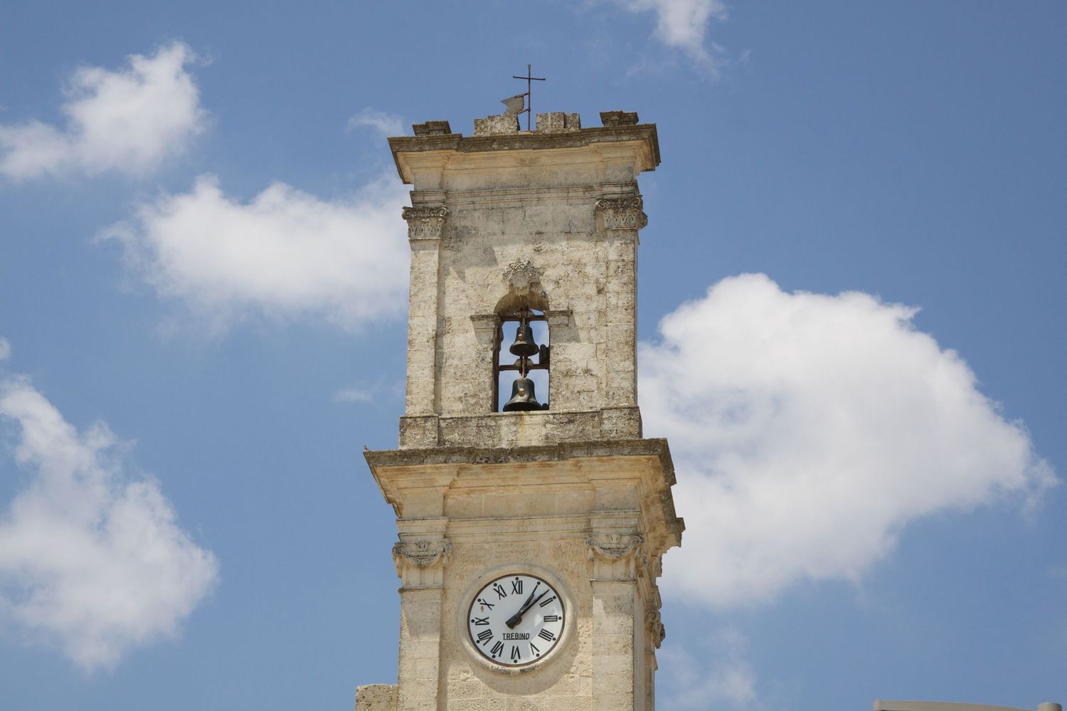 Historic stone bell tower with clock face and cross atop against blue cloudy sky.
