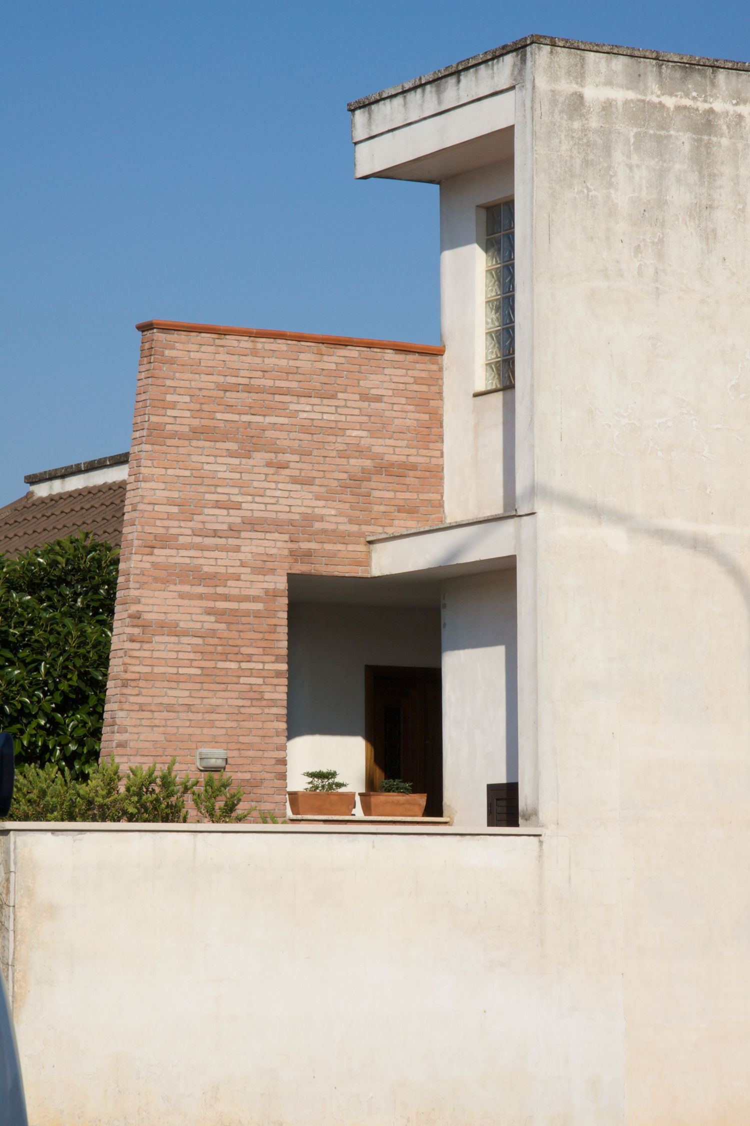 Modern residential building combining white render and red brick facade with balcony and potted plants against blue sky.