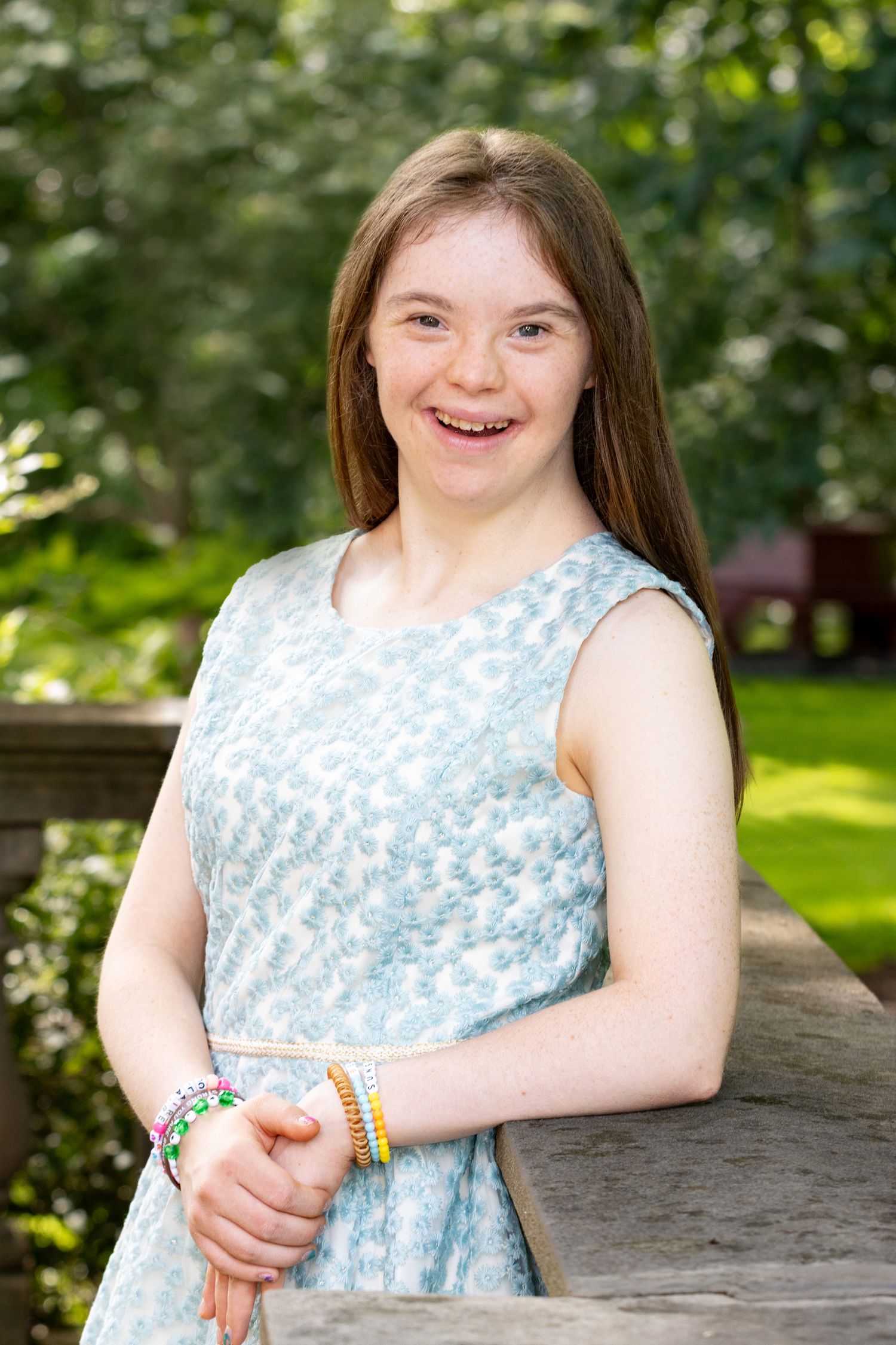 Smiling teenage girl in light blue dress wearing colorful bracelets outdoors.