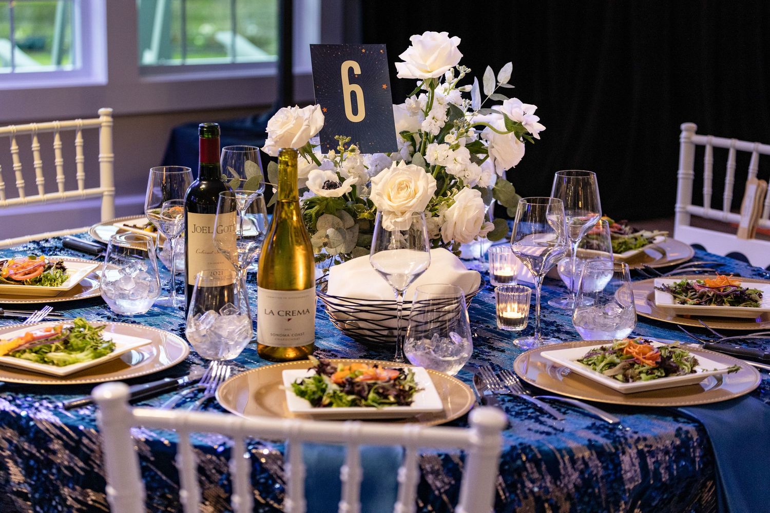 Elegant table setting with white roses, wine, and blue sequin tablecloth at a formal dinner.