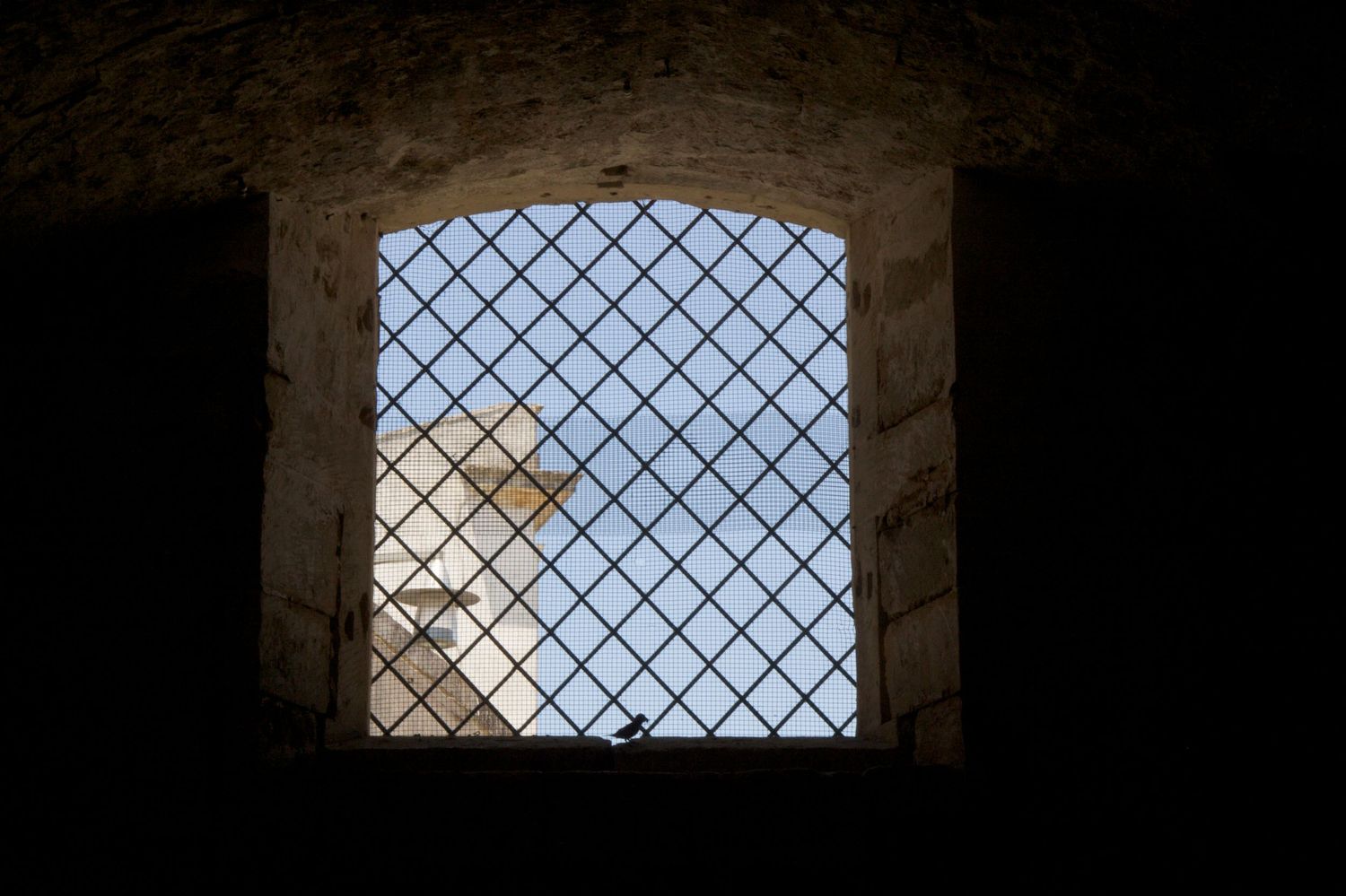 View through dark stone window with diamond iron grille revealing ancient stone tower or fortress in daylight.