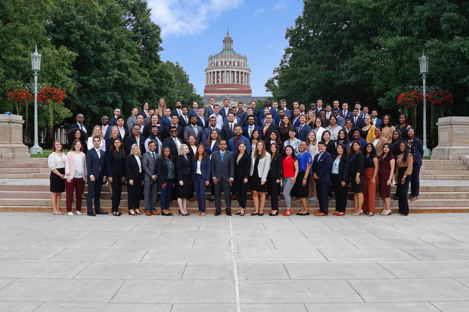 Large group of professionally dressed students pose on university steps with domed building backdrop.
