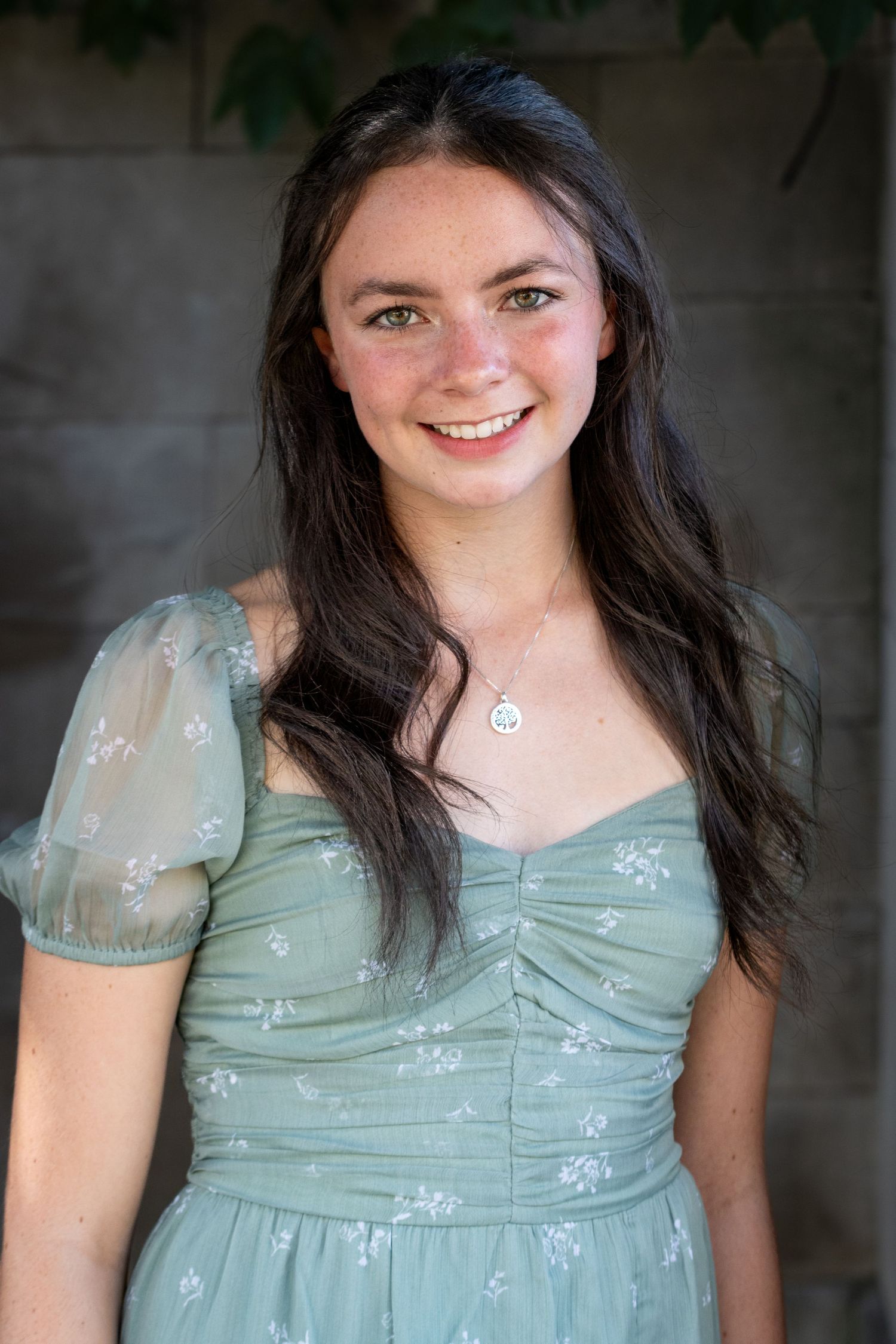 Young woman smiling in sage green floral dress with delicate necklace outdoors.
