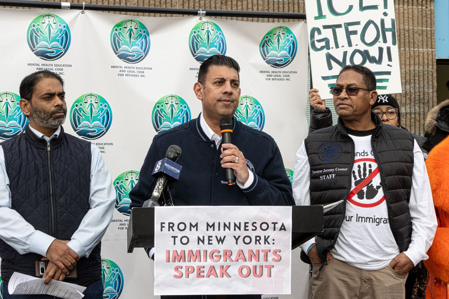 Man speaking at an immigrants rights rally with protesters holding anti-ICE signs behind him.