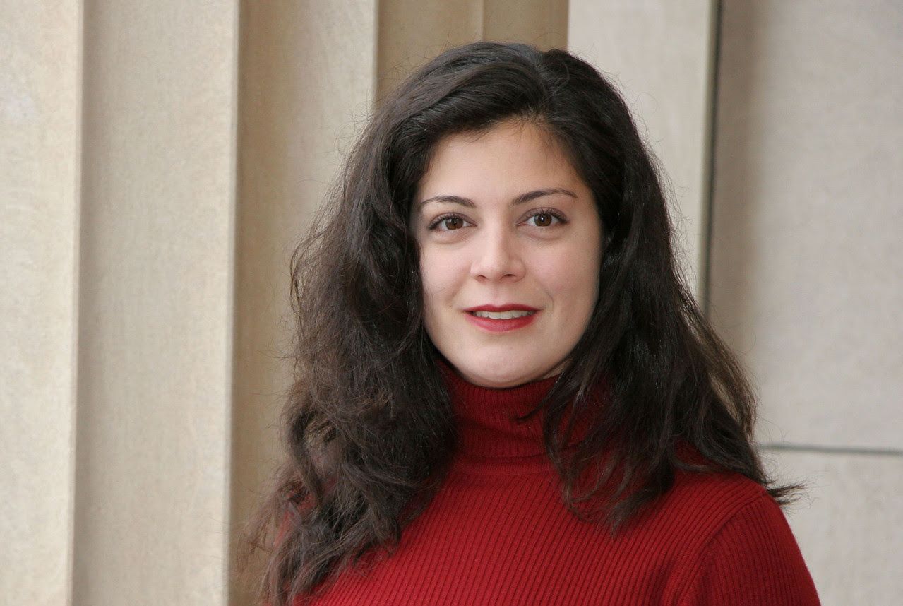 Young woman with dark curly hair in red turtleneck posing beside stone columns.