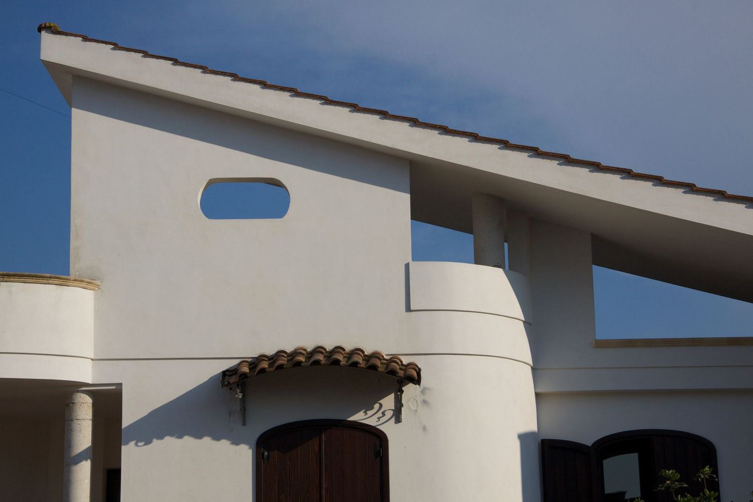 White Mediterranean-style villa with cylindrical columns, terracotta roof tiles and oval window detail against blue sky.