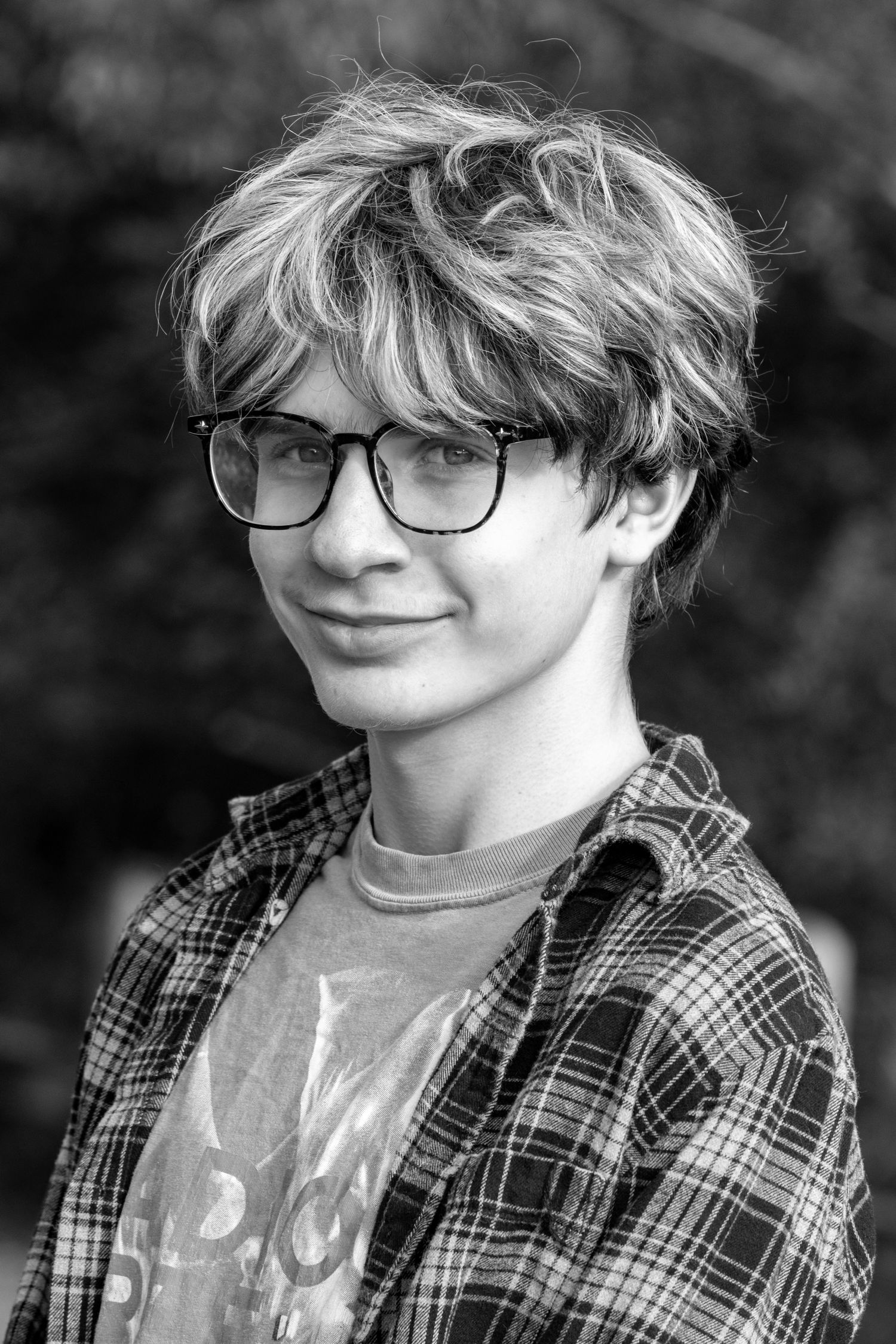 Black and white portrait of a teenage boy with wavy hair and glasses wearing a plaid shirt outdoors.