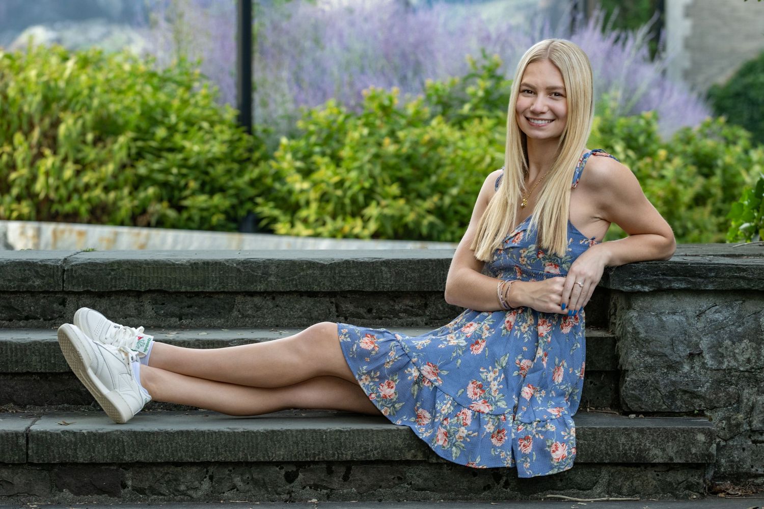 Blonde woman in floral blue dress sitting on stone steps surrounded by lush green garden.