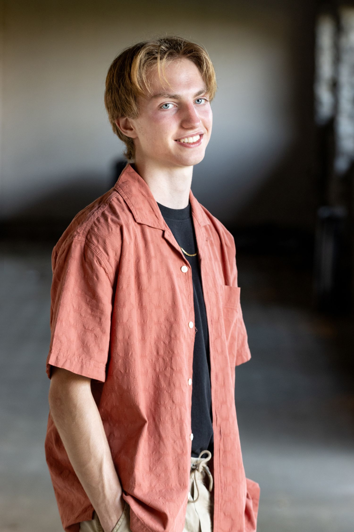 Smiling teenage boy in coral button-down shirt standing in dimly lit space.