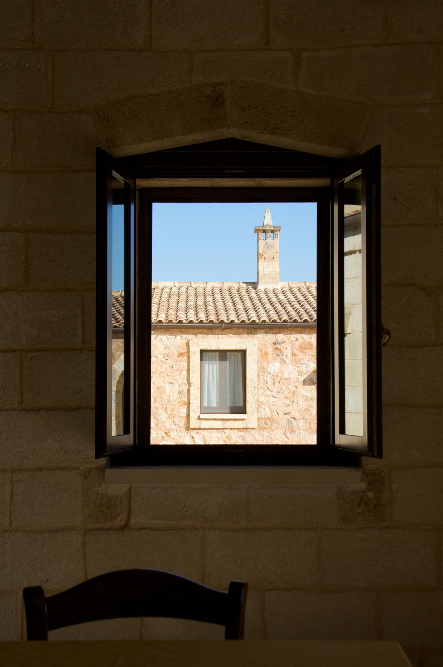 Open wooden window shutters revealing a sunlit stone building with chimney and tiled roof.