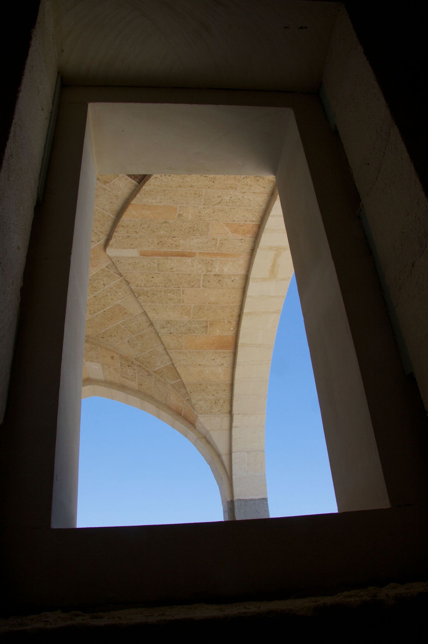 Low angle view looking up through a stone archway framing vaulted limestone arches against a clear blue sky.