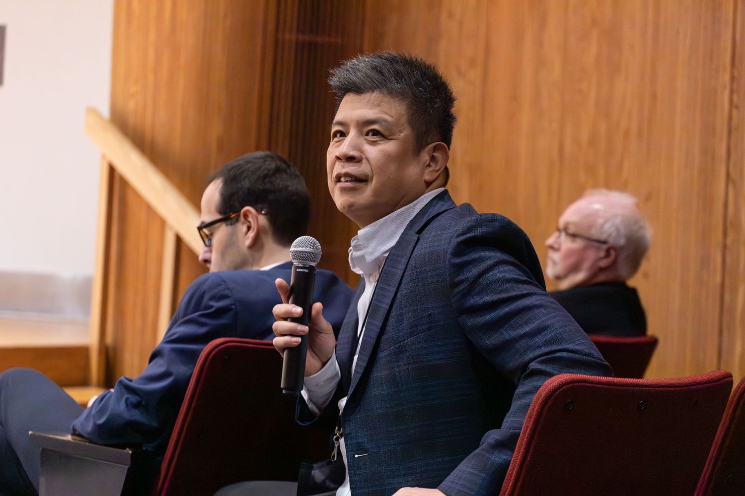 Man in blue blazer speaking into a microphone at a formal conference or panel discussion.