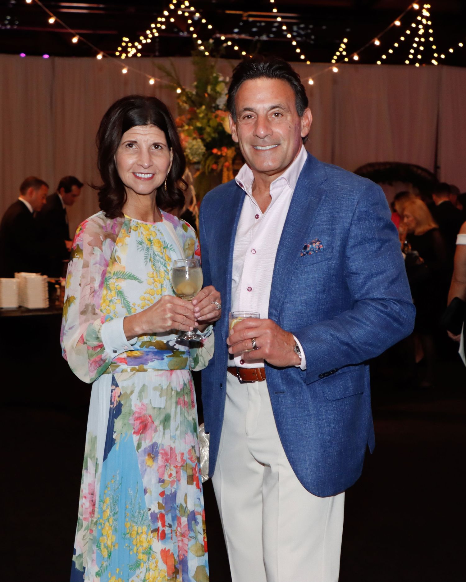 Elegant couple posing at formal gala event, woman in floral dress holding wine, man in blue blazer.