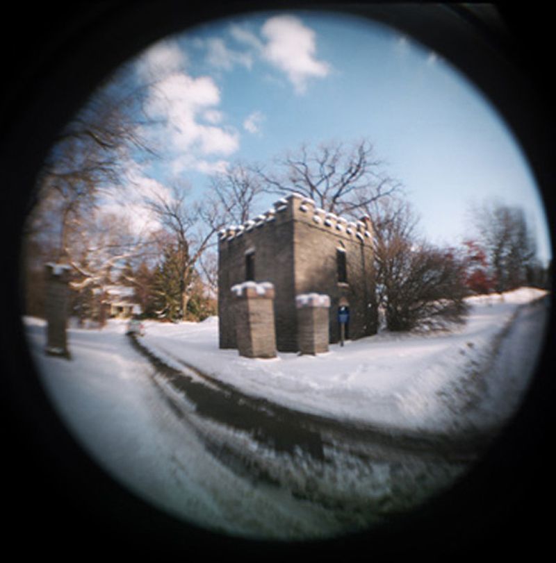 Fisheye lens photo of a small stone castle gatehouse along a snowy road lined with bare trees in winter.