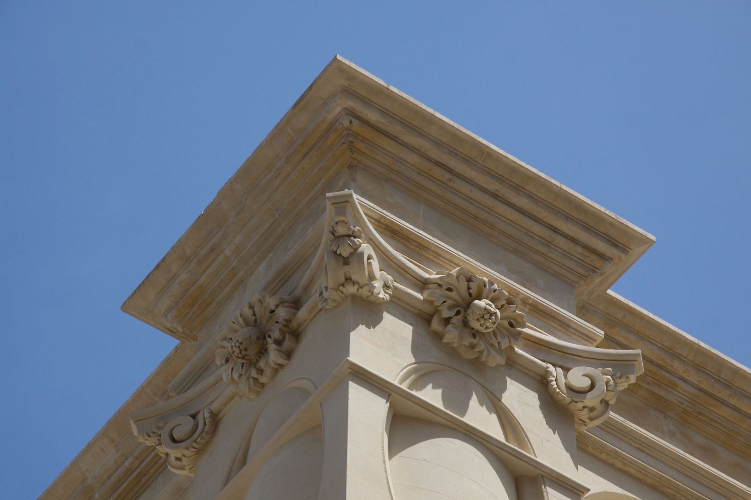 Ornate classical stone cornice with floral carvings and decorative moldings against a clear blue sky.