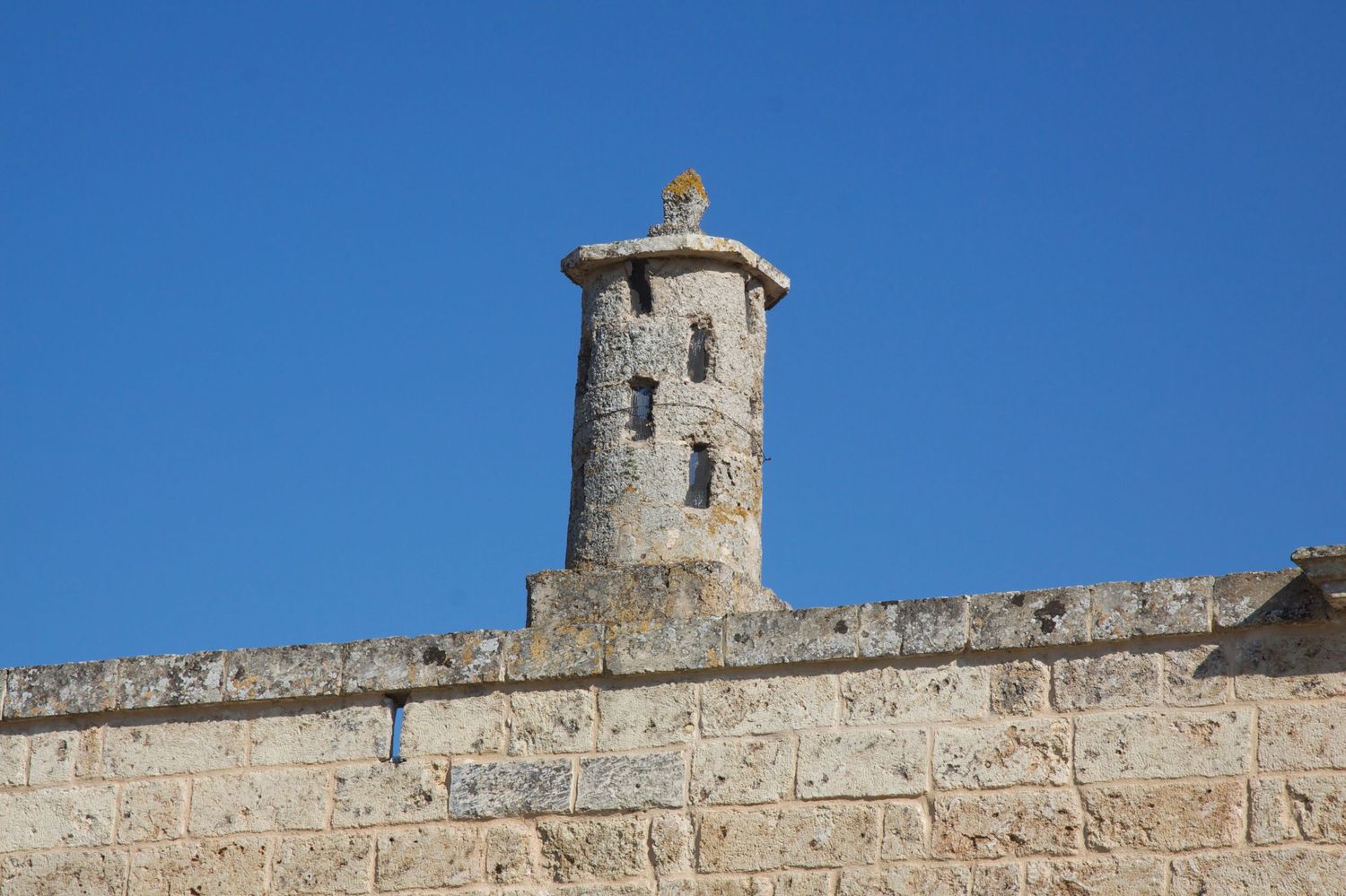 Ancient stone watchtower turret rising above a historic fortress wall against a clear blue sky.