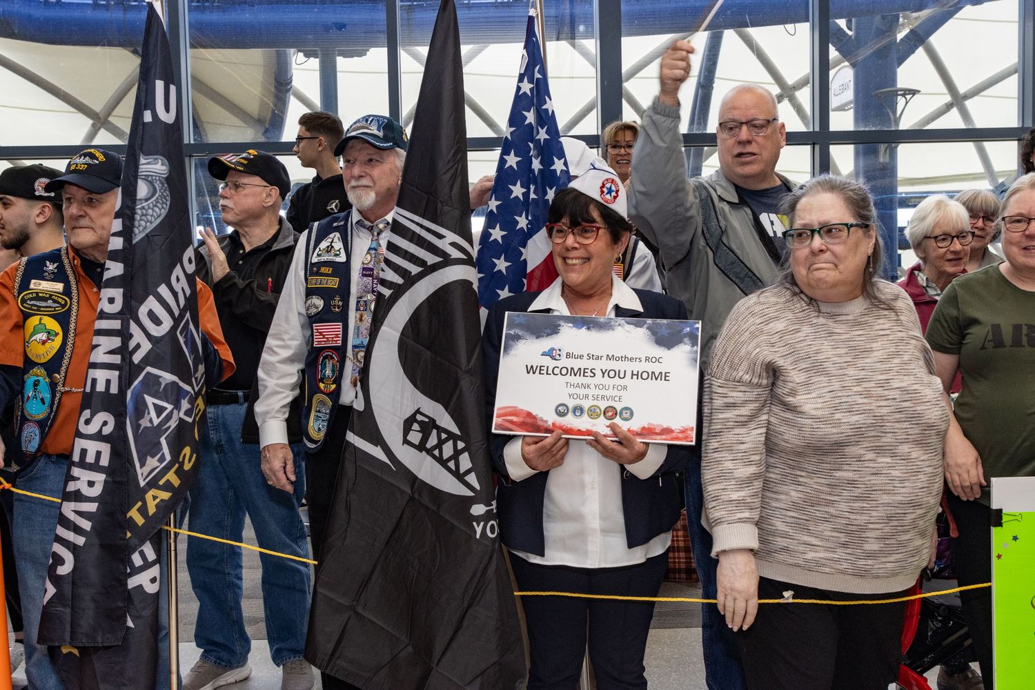 Veterans and supporters holding flags and welcome signs at a patriotic indoor ceremony.