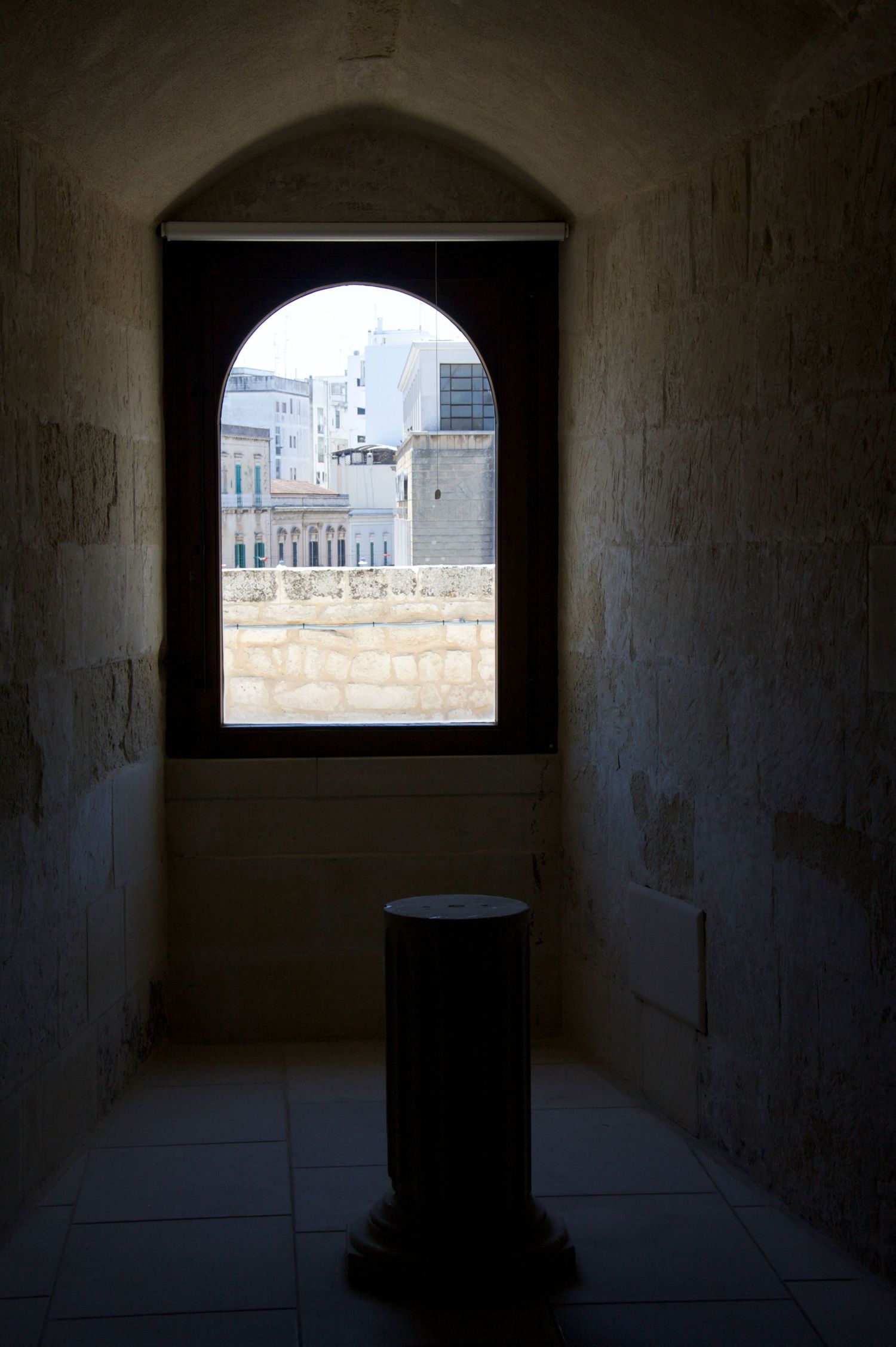 Ancient stone alcove with arched window overlooking modern city buildings, featuring a stone pedestal in dim interior light.