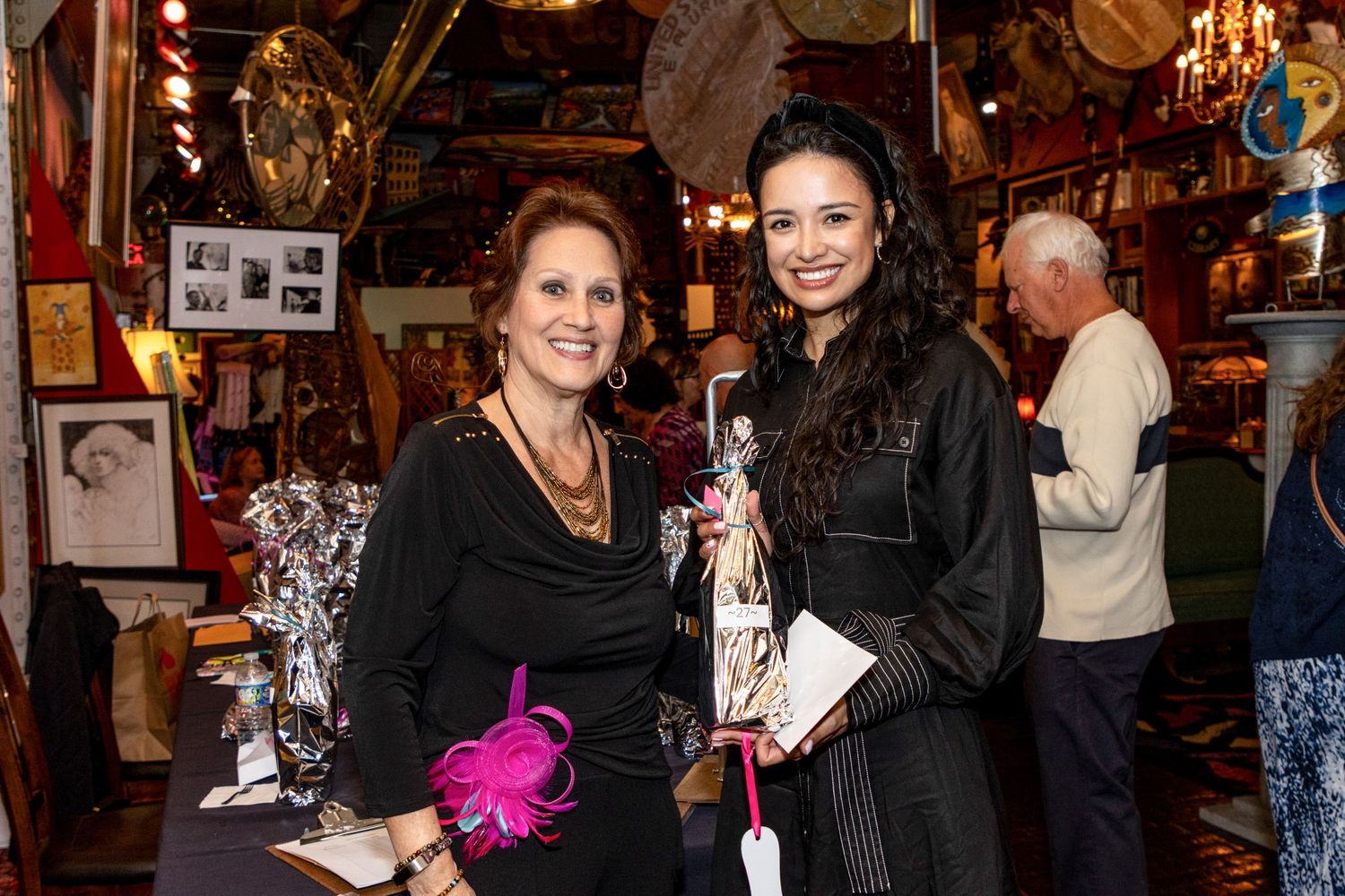 Two elegantly dressed women smile and pose together at an indoor event, one holding a decorative gift bag.