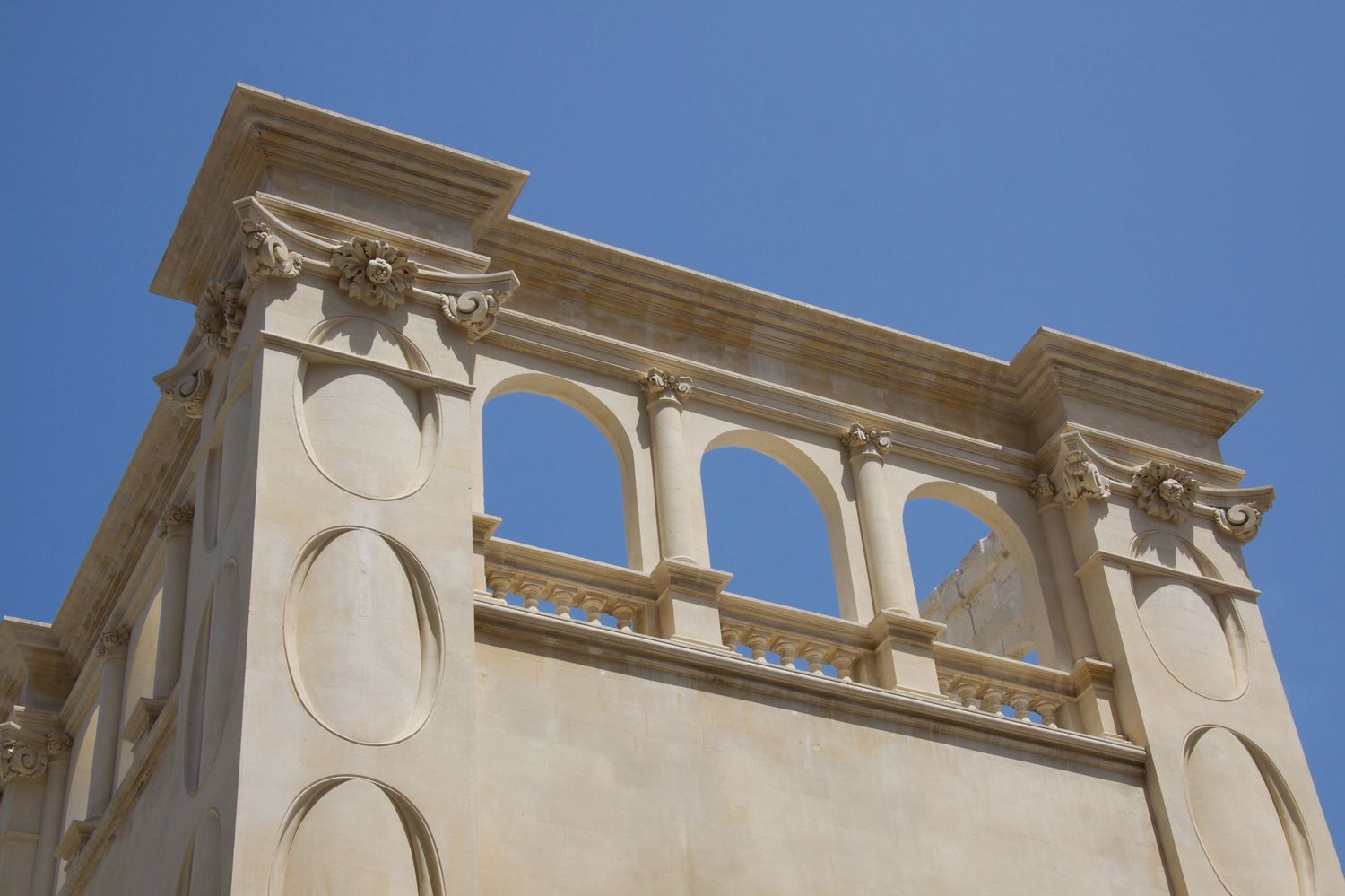 Ornate classical building top with arched balcony, decorative columns, and carved details against blue sky.