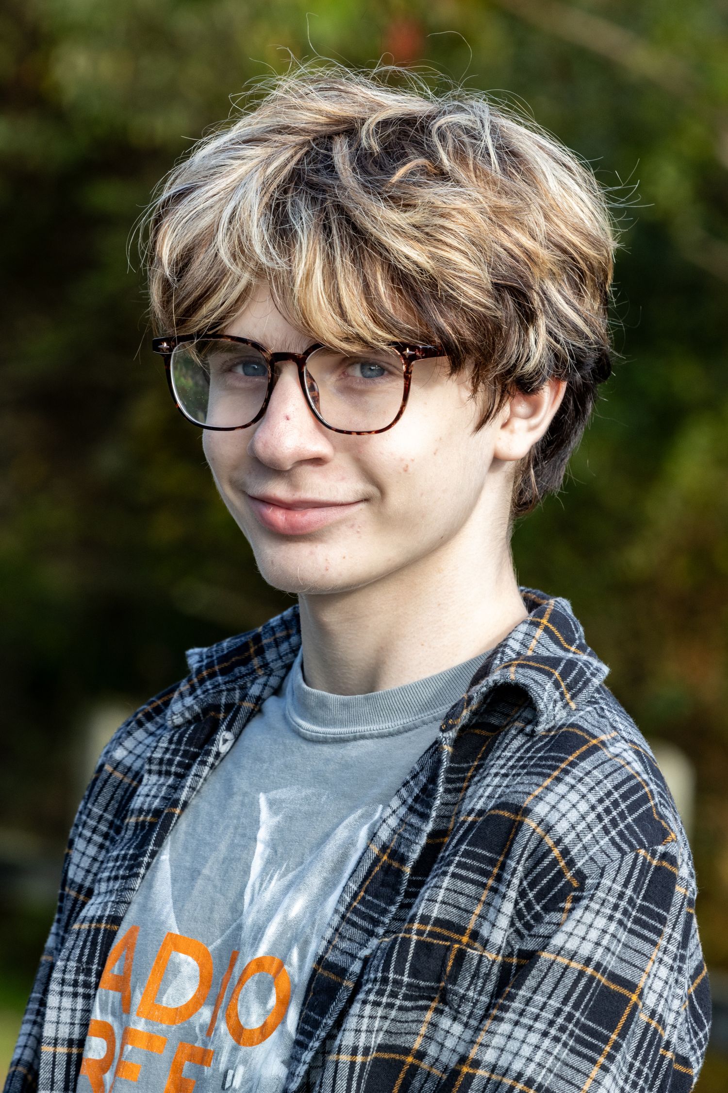 Teenage boy with glasses and plaid shirt smiling softly in an outdoor autumn setting.