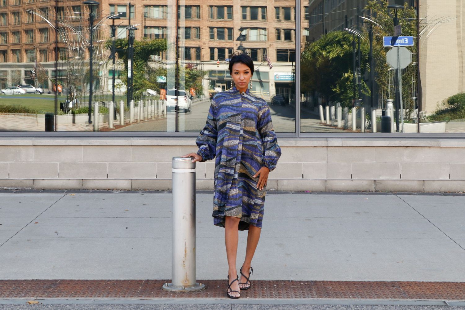 Woman in blue camo dress posing by a bollard on an urban sidewalk with city buildings in the background.