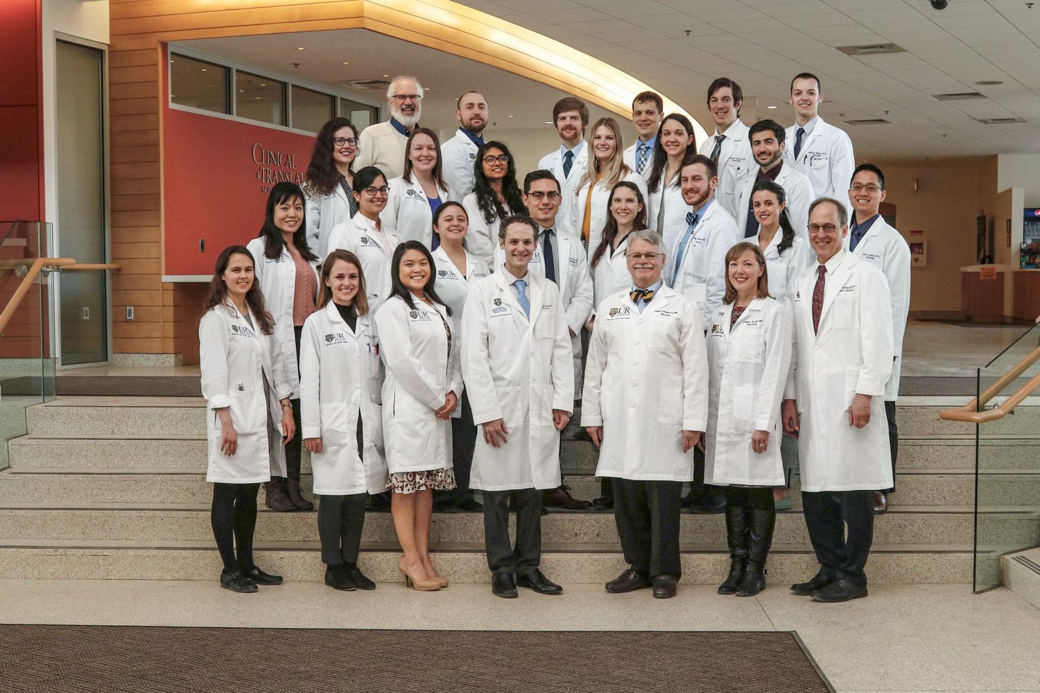 Group of medical professionals in white lab coats posing on stairs in a modern hospital atrium.